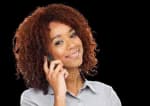 A woman with curly hair and a light gray shirt smiles while holding a smartphone to her ear against a plain background.
