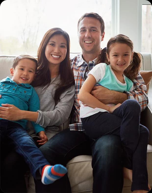 A family of four sits on a couch, smiling at the camera. The mother and father each hold one child on their laps.