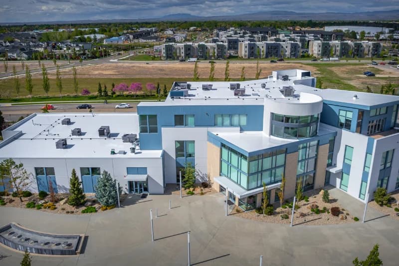 A modern, multi-story commercial building with a flat white roof, large glass windows, and landscaped surroundings, viewed from above with apartments and mountains in the background.