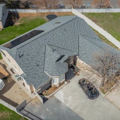 Aerial view of a beige two-story home with a light blue-gray shingle roof featuring solar panels and a small turret-style entry.