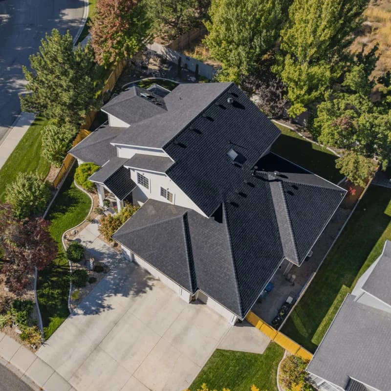 Aerial view of a white two-story house with a sharp black shingle roof, manicured landscaping, and tree-lined borders.