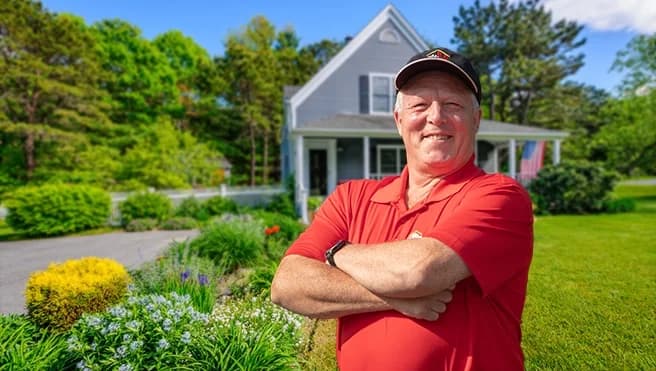 Smiling male handyman in red shirt standing with arms crossed in front of residential home with landscaped driveway.