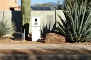 A white utility box and large cactus in a desert landscape, with a solar panel on a nearby wall.