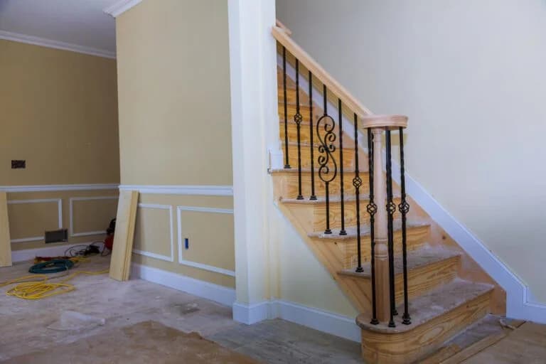Staircase with wooden steps and decorative black iron railing in a partially finished room, showcasing custom cabinetry and trim work by Ace Kustoms.