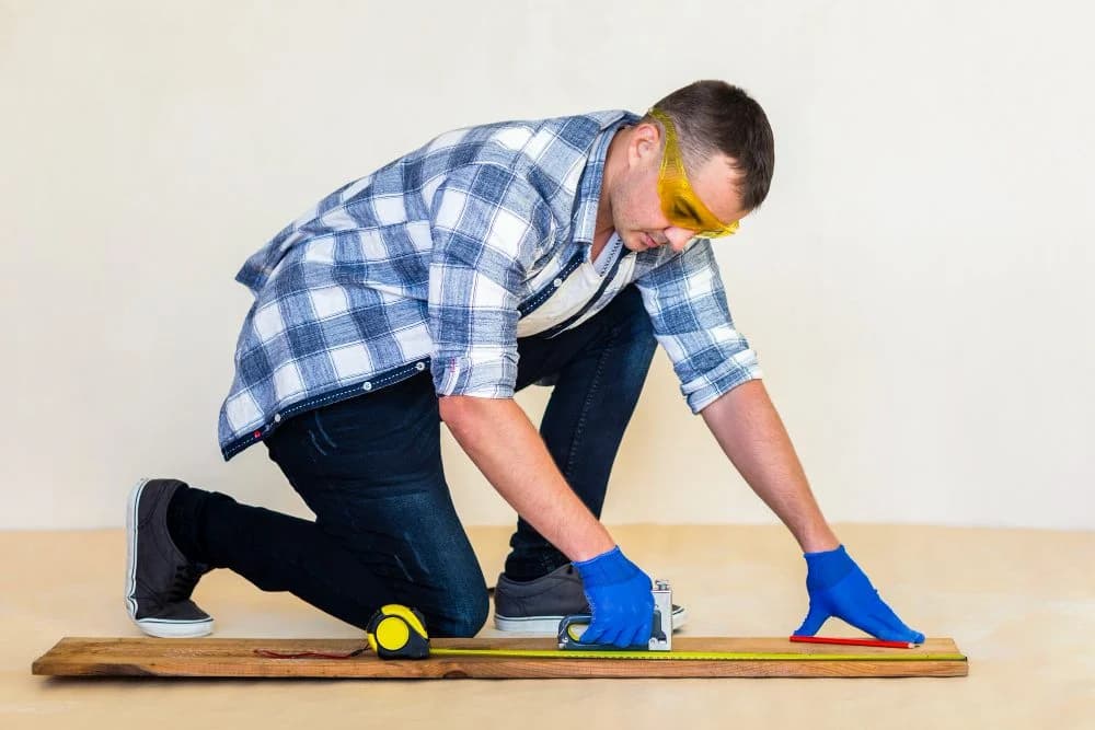 Man measuring wood with a tape measure and square, wearing safety goggles and gloves, illustrating craftsmanship in custom trim work.