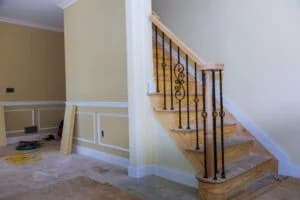 Staircase with wooden steps and decorative black iron railing in a partially finished room, showcasing custom cabinetry and trim work by Ace Kustoms.