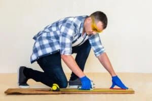 Man measuring wood with a tape measure and square, wearing safety goggles and gloves, illustrating craftsmanship in custom trim work.
