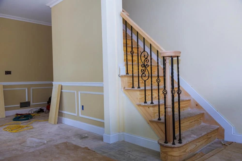 Staircase with wooden steps and decorative black iron railing in a partially finished room, showcasing custom cabinetry and trim work by Ace Kustoms.