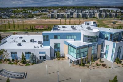 A modern, multi-story commercial building with a flat white roof, large glass windows, and landscaped surroundings, viewed from above with apartments and mountains in the background.