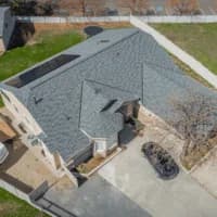Aerial view of a beige two-story home with a light blue-gray shingle roof featuring solar panels and a small turret-style entry.
