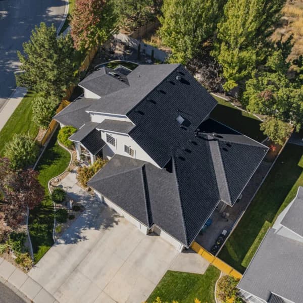 Aerial view of a white two-story house with a sharp black shingle roof, manicured landscaping, and tree-lined borders.