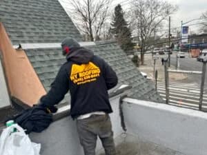 A person wearing a NY Roofing jacket stands on a rooftop near a sloped roof and looks down, with a busy street and leafless trees visible in the background.