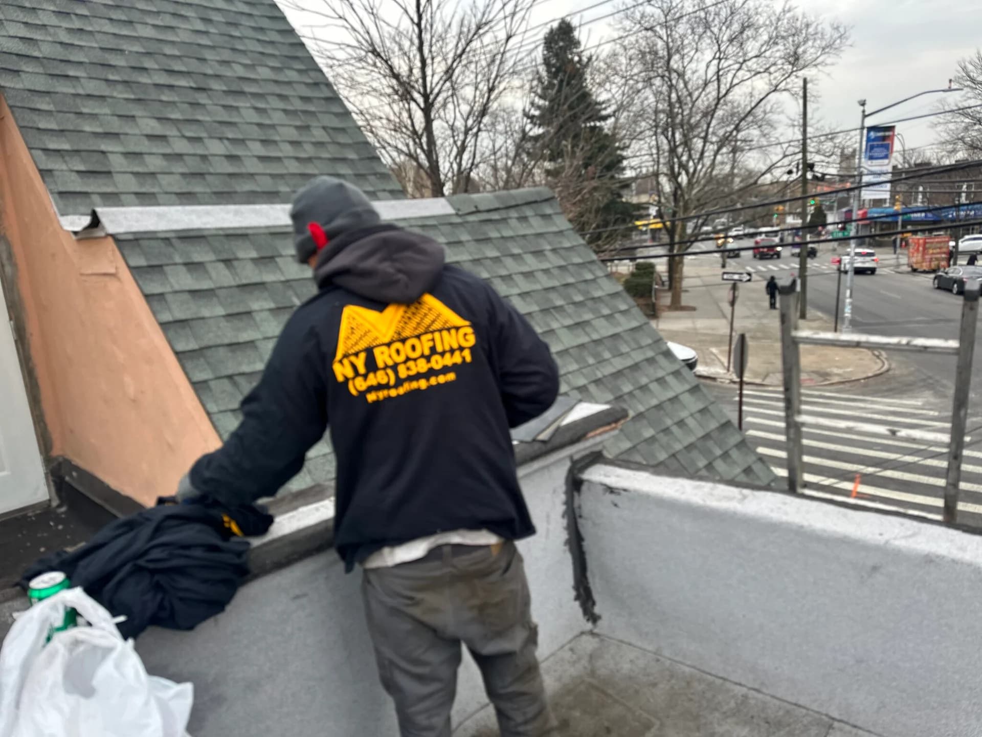 A person wearing a NY Roofing jacket stands on a rooftop near a sloped roof and looks down, with a busy street and leafless trees visible in the background.