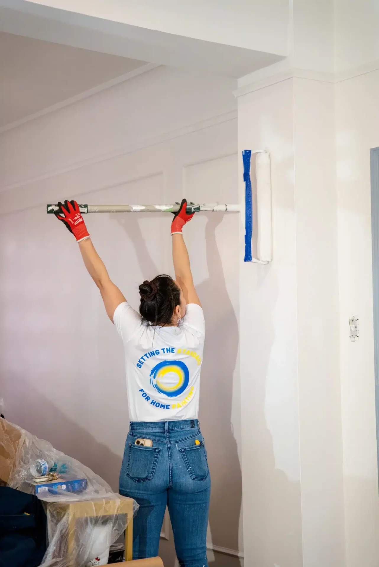 Professional painter in company uniform applying paint to a white wall and ceiling with an extension roller.