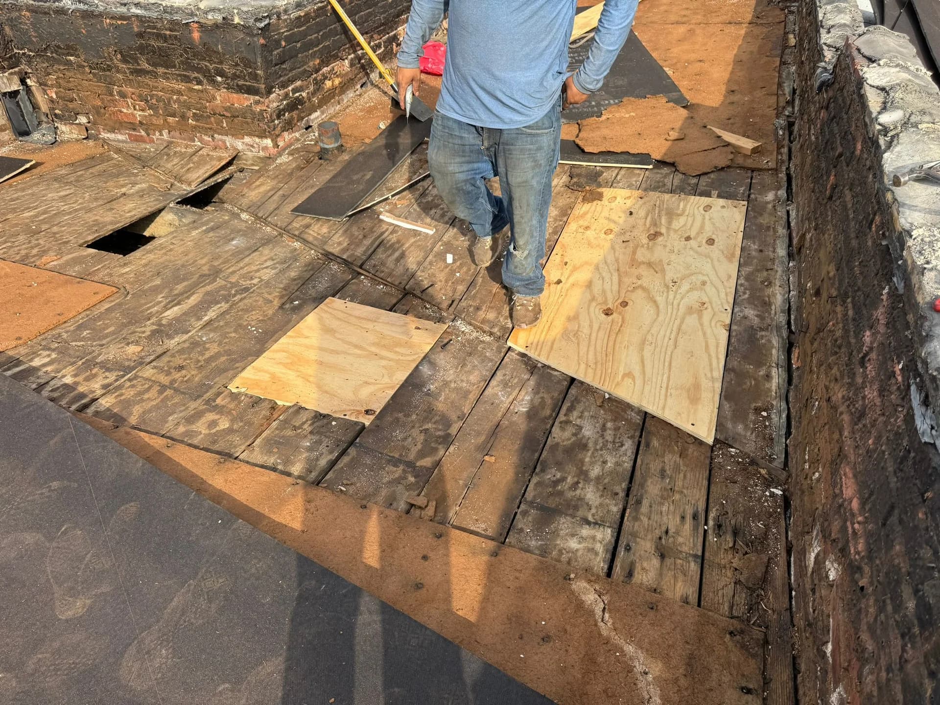 Worker installing plywood on a flat roof deck during rubber roof replacement, with visible areas of old roofing material and preparation for new EPDM membrane.