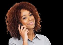 A woman with curly hair and a light gray shirt smiles while holding a smartphone to her ear against a plain background.