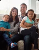 A family of four sits on a couch, smiling at the camera. The mother and father each hold one child on their laps.