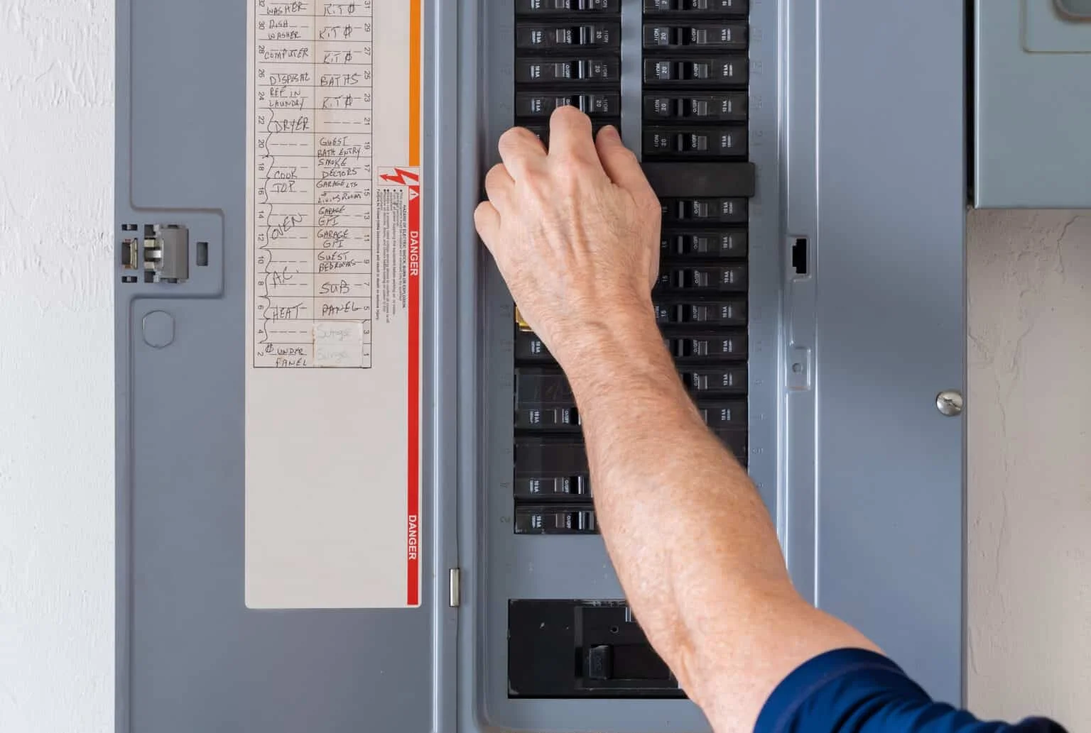 A person’s hand is adjusting a switch inside an electrical breaker panel, with a list of circuit labels visible on the panel door.
