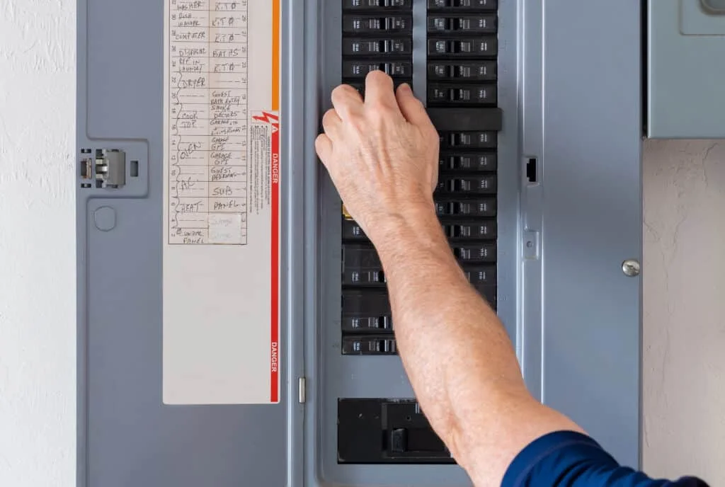 A person’s hand is adjusting a switch inside an electrical breaker panel, with a list of circuit labels visible on the panel door.