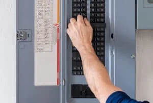 A person’s hand is adjusting a switch inside an electrical breaker panel, with a list of circuit labels visible on the panel door.