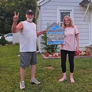 Two people standing in front of a small house during sunset holding a Great Lakes Home Remodeling sign