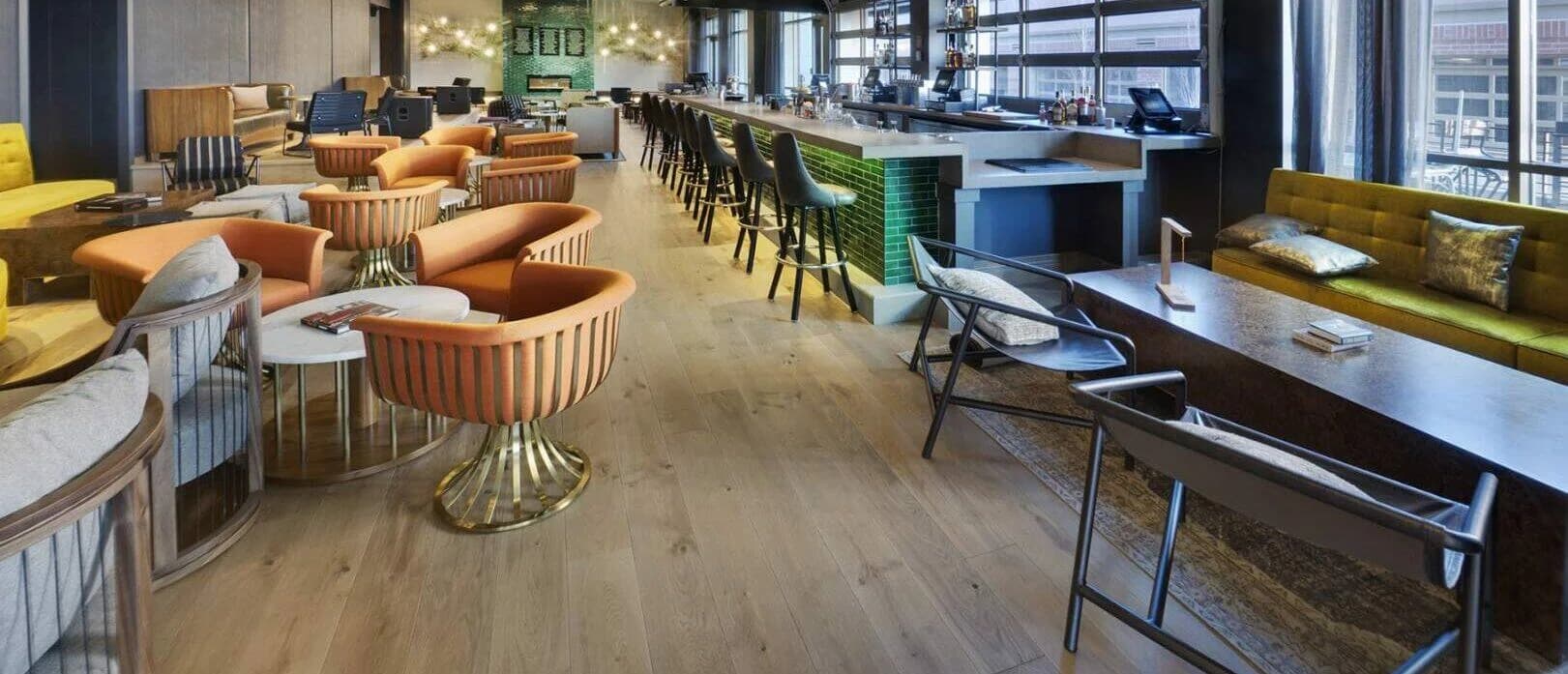 A wide shot of a long room with a well-lit bar, many options of comfortable seating, and a commercially done wooden floor