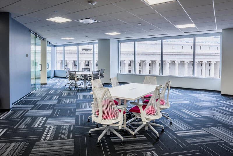 Modern office meeting area with white table, ergonomic chairs, and large windows showing a stone building with columns.