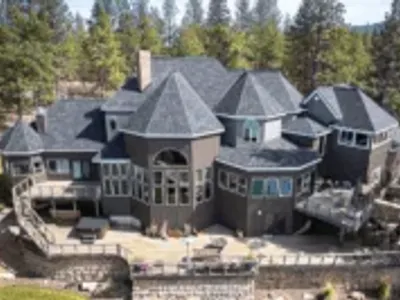 A large, multi-level home with dark gray asphalt shingles, multiple gables, and a spacious wraparound deck, surrounded by tall pine trees.