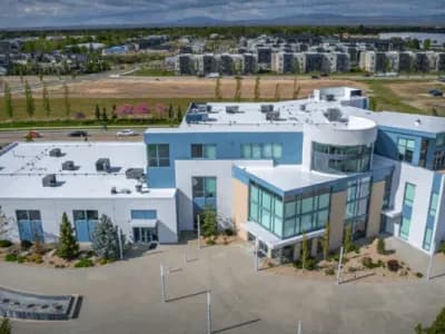 A modern, multi-story commercial building with a flat white roof, large glass windows, and landscaped surroundings, viewed from above with apartments and mountains in the background.