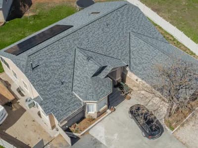 Aerial view of a beige two-story home with a light blue-gray shingle roof featuring solar panels and a small turret-style entry.
