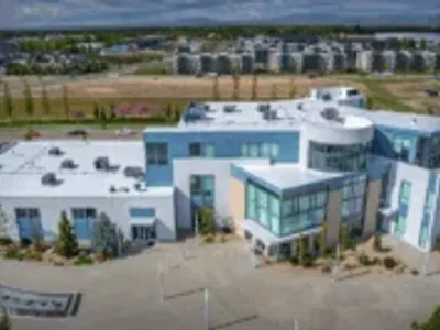 A modern, multi-story commercial building with a flat white roof, large glass windows, and landscaped surroundings, viewed from above with apartments and mountains in the background.