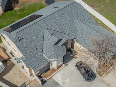 Aerial view of a beige two-story home with a light blue-gray shingle roof featuring solar panels and a small turret-style entry.