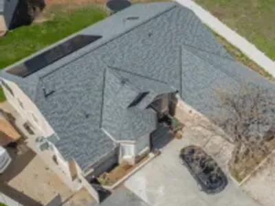 Aerial view of a beige two-story home with a light blue-gray shingle roof featuring solar panels and a small turret-style entry.