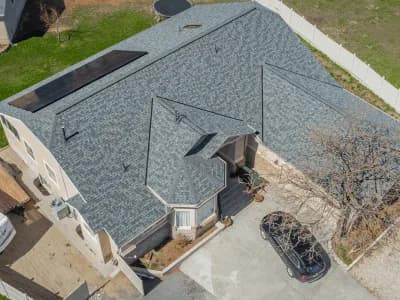 Aerial view of a beige two-story home with a light blue-gray shingle roof featuring solar panels and a small turret-style entry.