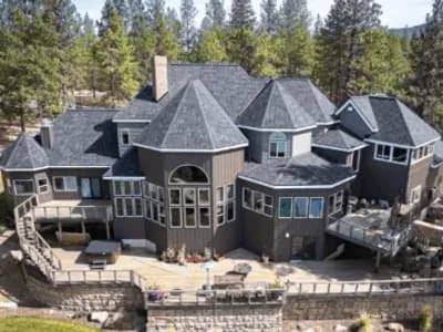 A large, multi-level home with dark gray asphalt shingles, multiple gables, and a spacious wraparound deck, surrounded by tall pine trees.
