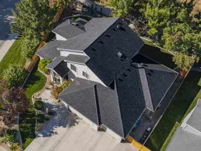 Aerial view of a white two-story house with a sharp black shingle roof, manicured landscaping, and tree-lined borders.