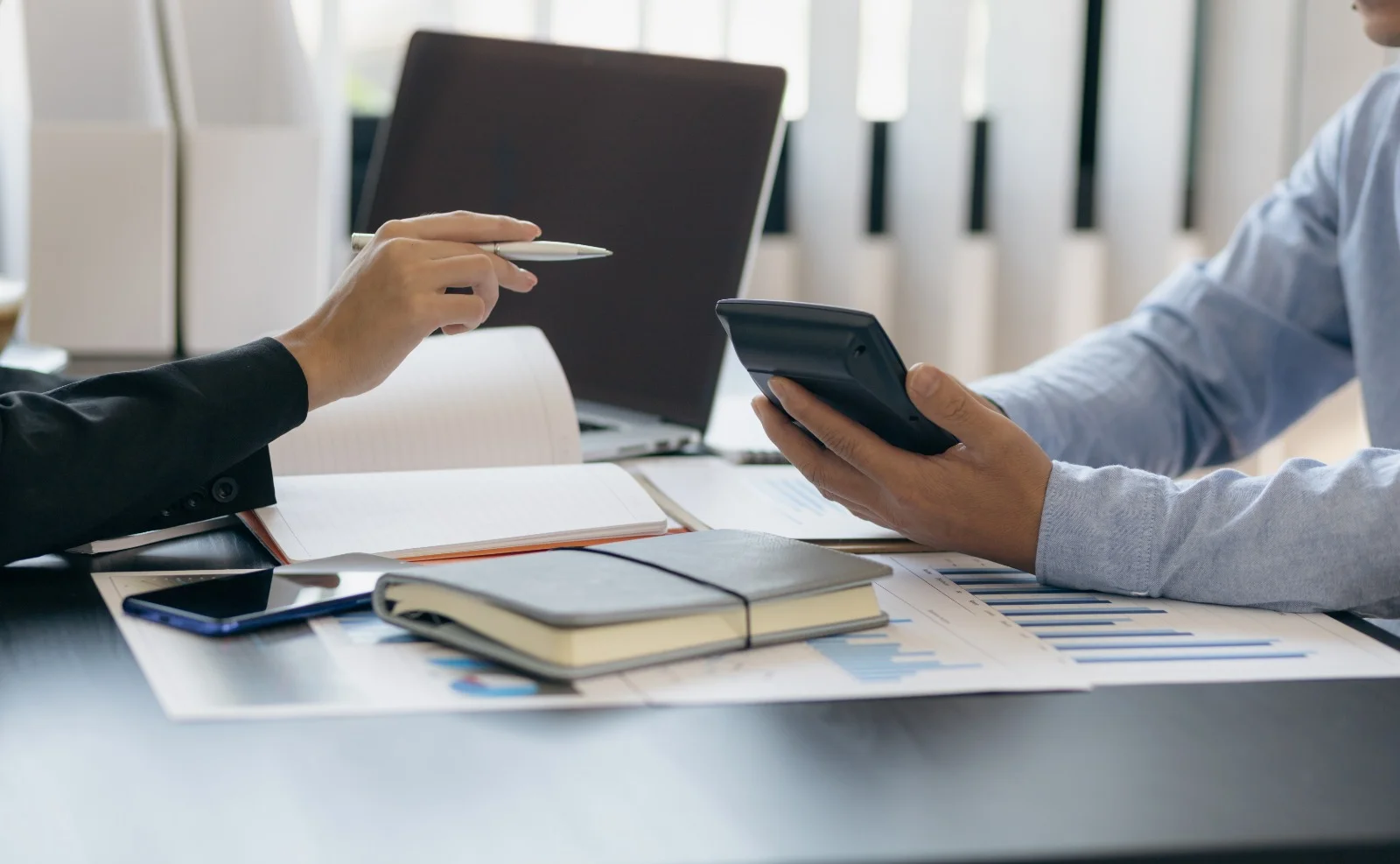 Two business professionals discussing financial reports at a desk with a calculator, notebooks, charts, laptop, and smartphone