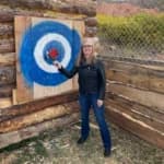Woman in a black leather jacket aiming a throwing axe at a target.