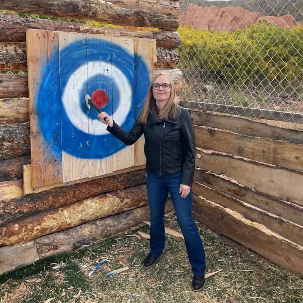 Woman in a black leather jacket aiming a throwing axe at a target.
