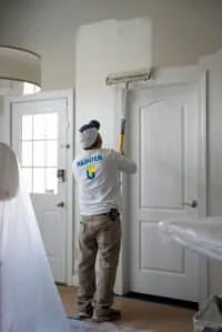 A professional painter applies fresh white paint to a residential interior wall above a door.
