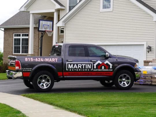 A black pickup truck with "Martin Exteriors" branding is parked on a driveway in front of a house. Contact information and logo are displayed on the truck.