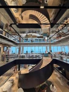 A modern, open office space features a dramatic spiral staircase with black railings and light wood steps. The staircase and workers are reflected in a large mirrored ceiling above.