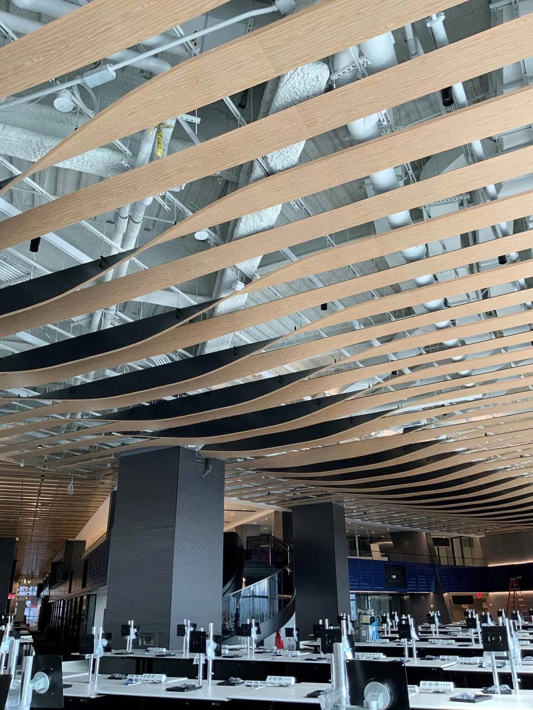 Curved wooden slats form a wave-like pattern on the ceiling of a modern, spacious interior with exposed ductwork above and tables and chairs below.