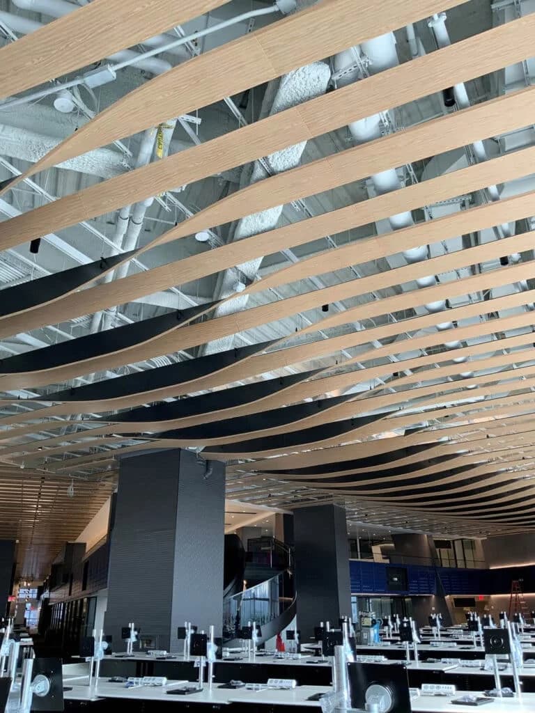 Curved wooden slats form a wave-like pattern on the ceiling of a modern, spacious interior with exposed ductwork above and tables and chairs below.