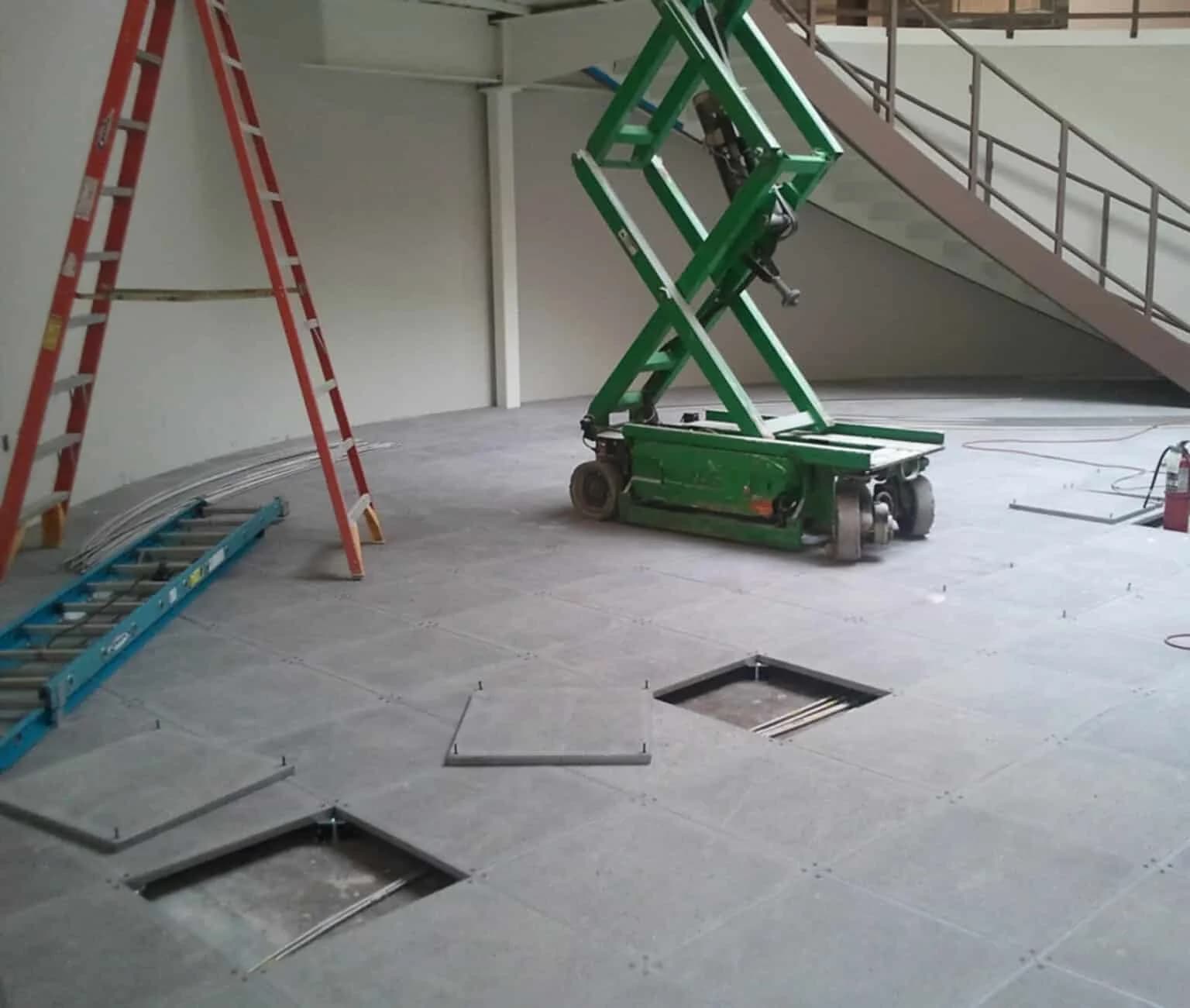 A room under construction with an open raised floor showing cables, a green scissor lift, two ladders (one red, one blue), and a staircase in the background. Some floor panels are removed.