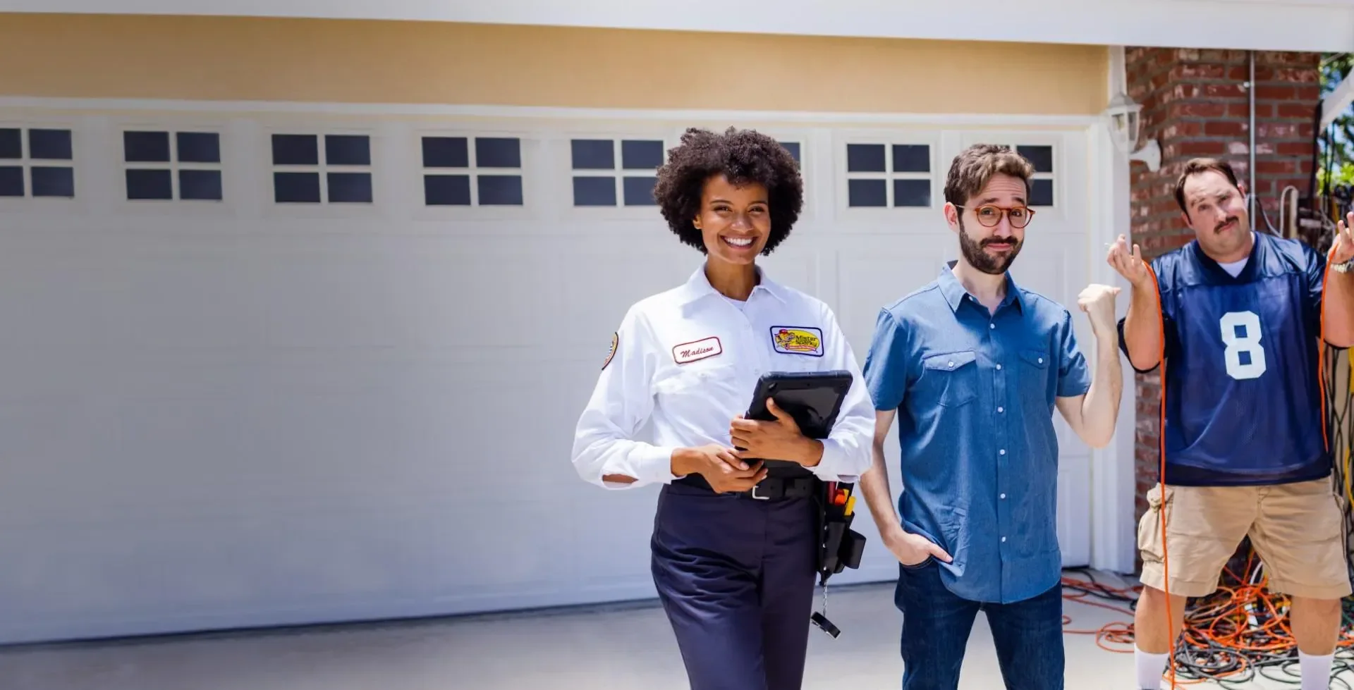 Female electrician with a tablet smiling at the camera next to two customers in front of a garage.